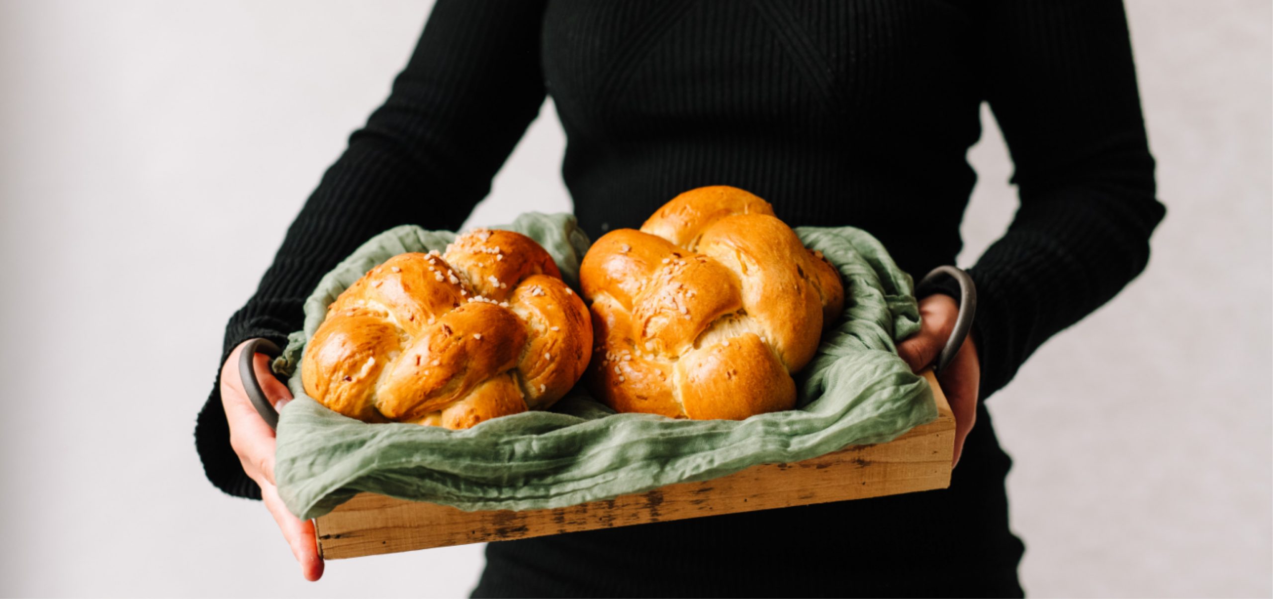 Eine Person hält ein Holztablett in die Kamera auf dem zwei Hefezöpfe auf einem grünen Tuch angerichtet wurden.