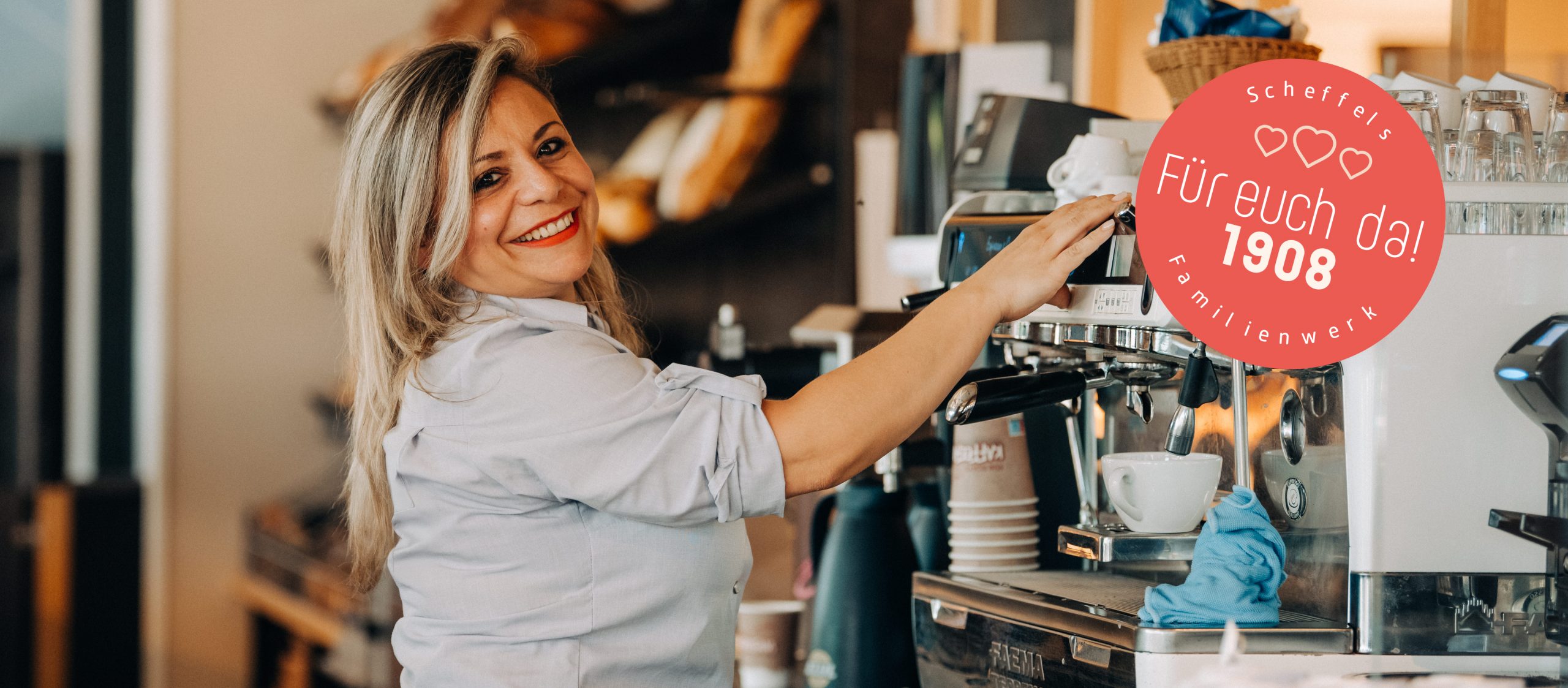 Eine Scheffels Mitarbeiterin steht an der Siebträgermaschine und lächelt in die Kamera. auf der rechen Seite ist ein roter Kreis auf das Bild gesetzt worden mit der Aufschrift Scheffels Familienwerk – Für euch da! 1908