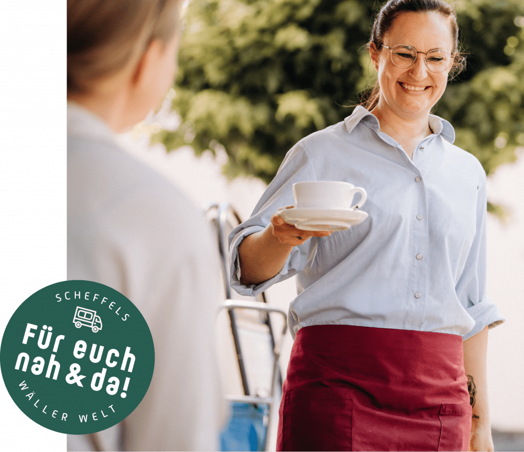 Ein Bild einer Verkäuferin der Bäckerei Scheffels, die gerade einer Kundin einen Kaffee zum Tisch bringt. Auf der linken Seite befindet sich ein Grüner Kreis mit der Aufschrift Scheffels Wäller Welt – Für euch nah und da!