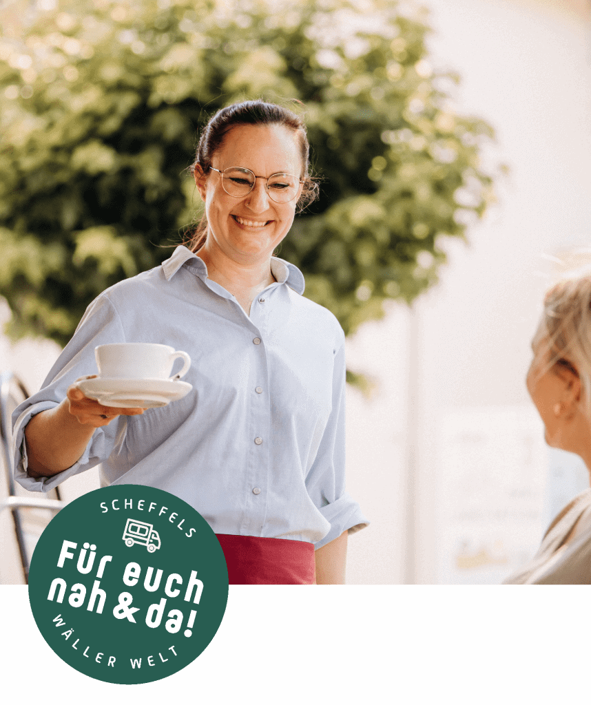 Ein Bild einer Verkäuferin der Bäckerei Scheffels, die gerade einer Kundin einen Kaffee zum Tisch bringt. Auf der linken Seite befindet sich ein Grüner Kreis mit der Aufschrift Scheffels Wäller Welt – Für euch nah und da!